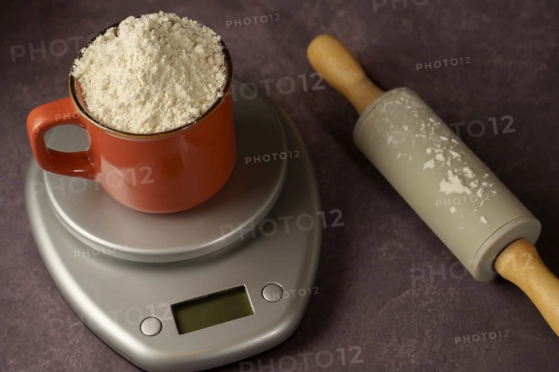 Red cup with flour on a kitchen scale next to a rolling pin with a dark background and copy space