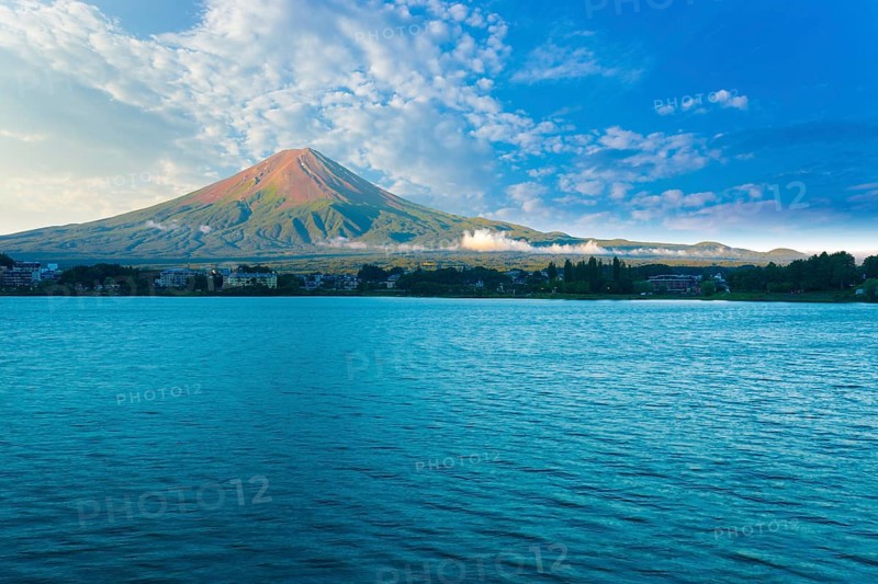 Mount Fuji bathes in morning side light while Lake Kawaguchiko sits in shadow below cloudy blue sky on a summer day in five lakes region of Japan. Horizontal