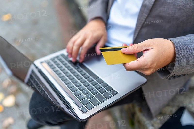 Close-up of a caucasian man shopping online using laptop and credit card sitting on city bench