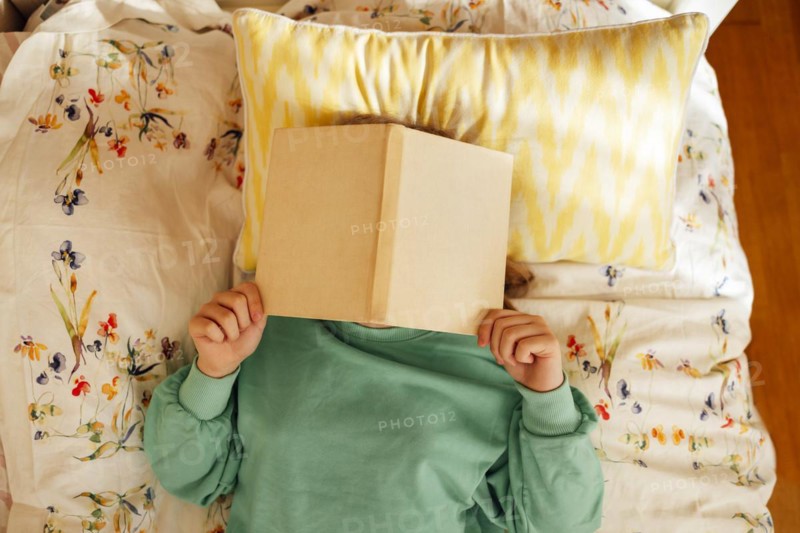 Caucasian blond teenage girl is lying on the bed and reading book. Reading fiction, hobbies, leisure activities and recreation. Concept of home education