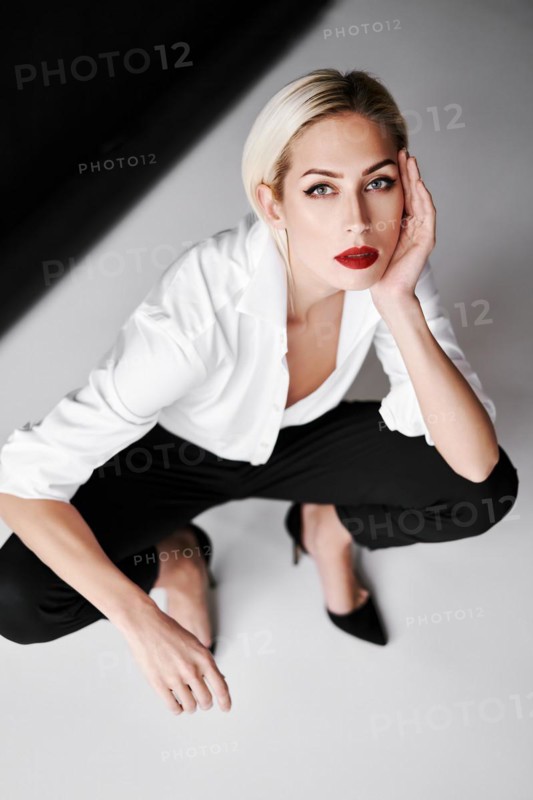 Top view portrait of daring trendy woman posing on white studio background sitting on floor. Gorgeous stylish blonde female wearing white shirt, black pants and high heel shoes sitting on floor