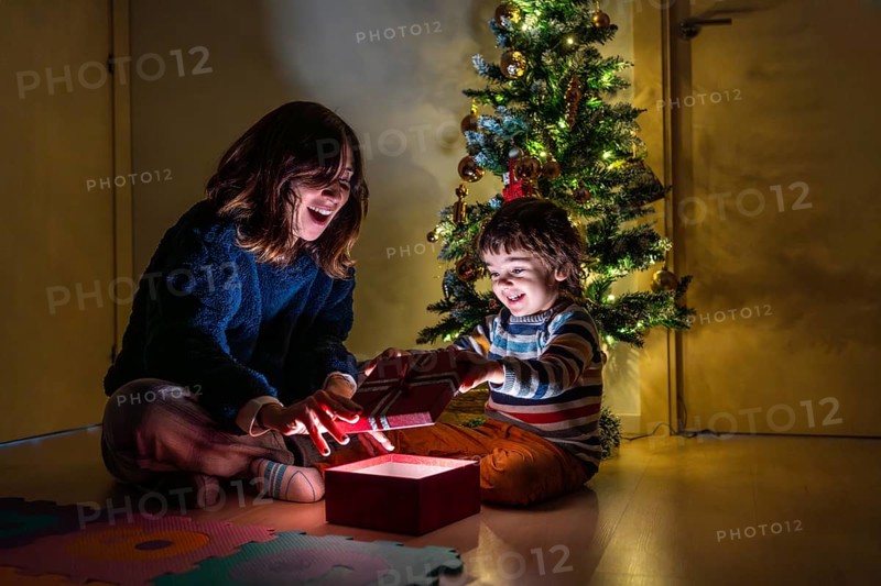 A young girl and boy are sitting on the floor in front of a Christmas tree. The girl is opening a present, and the boy is watching her. Scene is joyful and festive