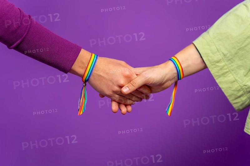 Studio photo with purple background and close-up of two lesbian woman holding hands wearing lgbt rainbow bracelet