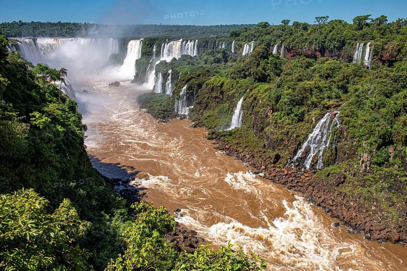View of Iguazu Falls gorge and Devil's Throat with brown river, white cascading falls and lush green rainforest in sunshine