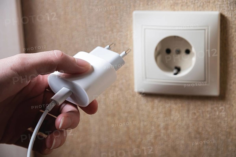 Hand turns on, turns off charger in electrical outlet on wall. White charger for phone in woman's hand close-up