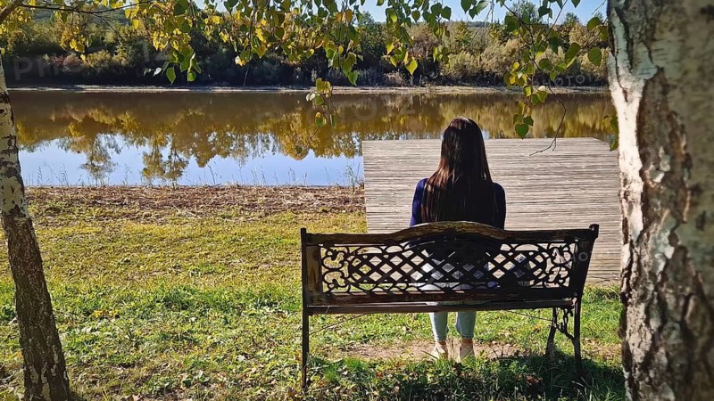 Single woman sitting on the bench at the lake surrounded by autumn foliage, beneath birch trees in the park. Peaceful fall season atmosphere