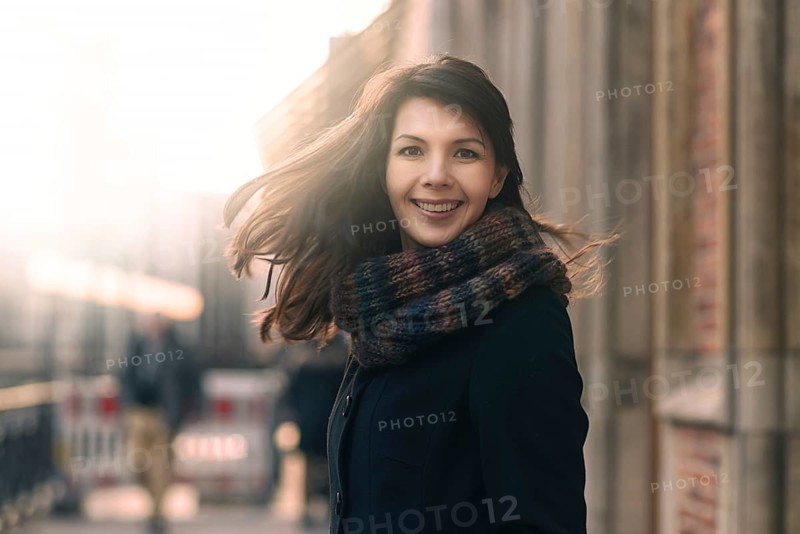 Happy woman with a lovely smile standing in her coat and scarf on an urban street in winter looking at the camera with a joyful expression and her hair blowing in the wind