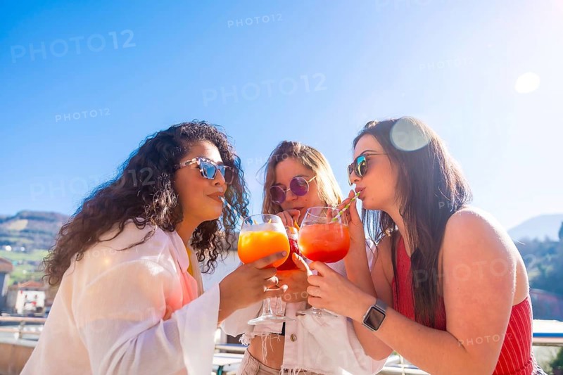 Women enjoying summer during a party drinking cocktails together with straw in a roof terrace