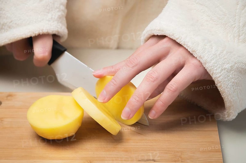 Young caucasian woman in a bathrobe stands in the kitchen and cuts fresh potatoes with a knife, on a wooden cutting board