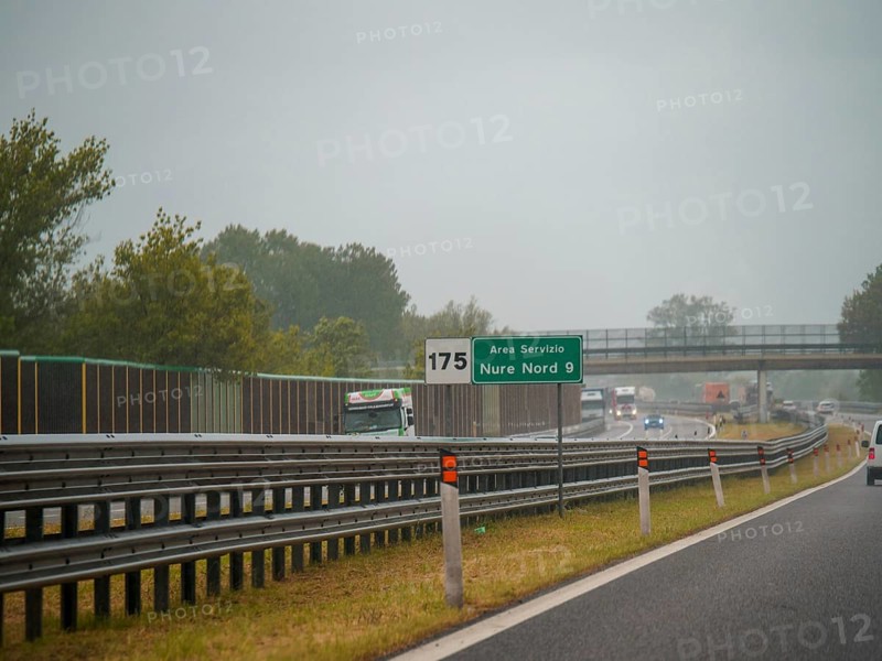 A highway scene under an overcast sky featuring cars and trucks with road signs and an overpass, rainy day on Motorway A1, Piacenza, Italy, Europe