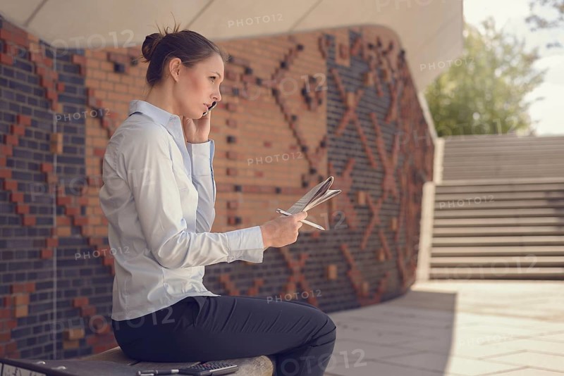 Woman doing business in a commercial foyer sitting on a wall mounted bench against patterned brickwork talking on her mobile phone discussing her notes with a tablet alongside, conceptual of mobility