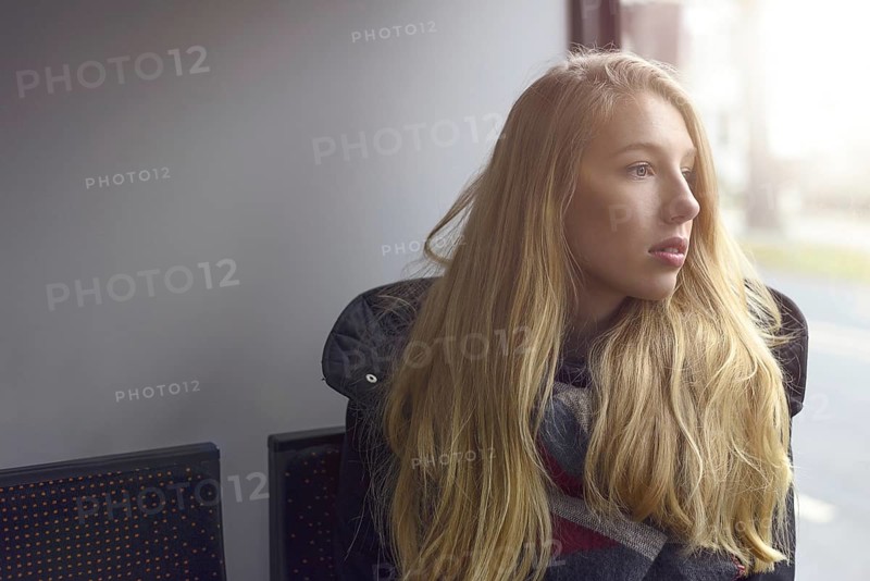 Young blond woman sitting in shadowed natural light looking out of a windows at the street outside with a serious thoughtful expression