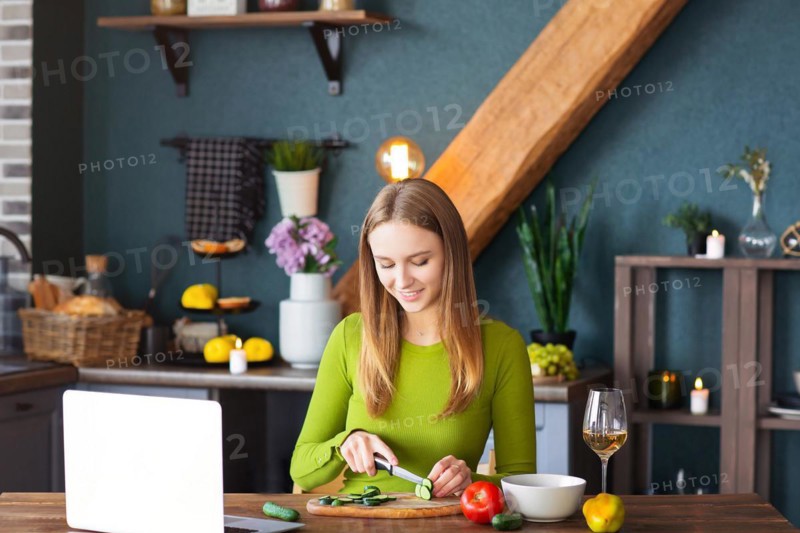 Happy female smiling and cutting cucumber for healthy vegetable salad while watching recipe on laptop in kitchen at home
