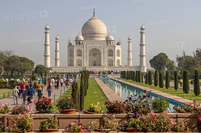 Taj Mahal, the mausoleum built for Mumtaz Mahal by her husband, the Mughal emperor, Shah Jahan. A distant, classic view, with some flowers in the foreground, the water channel reflecting the building, and some colourfully dressed visitors. Photographed in the morning of a mid-April day, the pre-monsoon summer season. Wonder of the World, a UNESCO World Heritage Site, famous landmark and tourist attraction. Agra, Uttar Pradesh, India, Asia