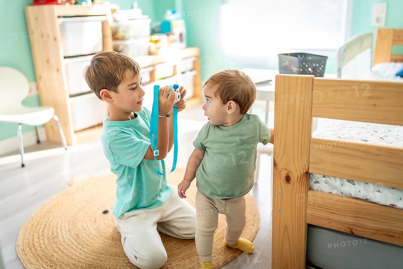 Male little photographer taking a photo of a his baby boy brother in the bedroom