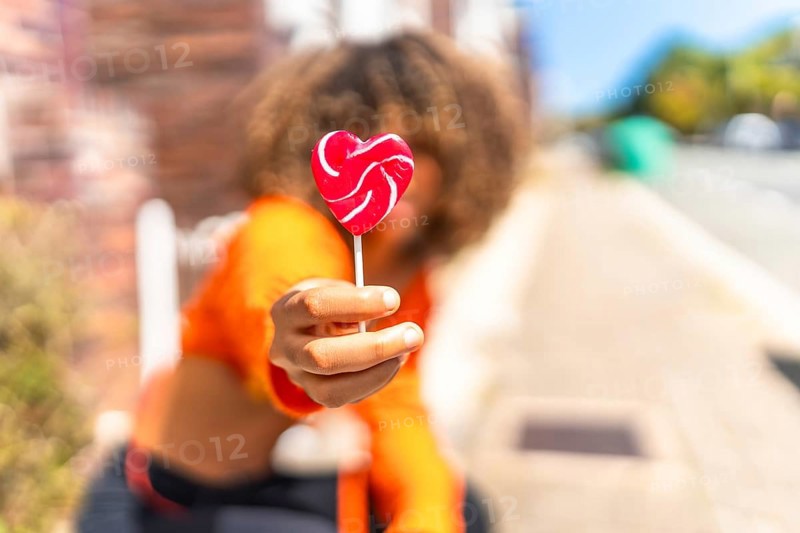 Focus on the hand of a young latin woman holding a heart shape lollipop in the street