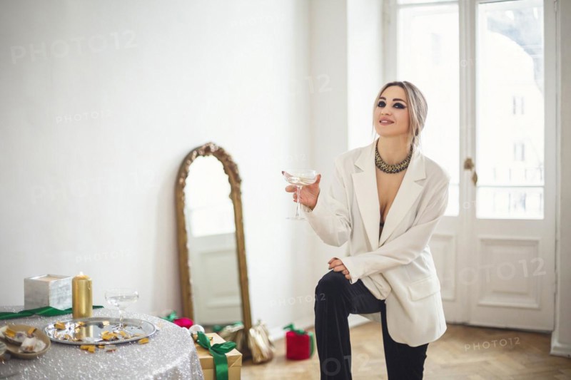 Young woman wearing sparkly sequin holiday dress sitting with alcohol drink in front of freestanding mirror in dressing room at home, female in festive wear getting ready for party in walk-in closet