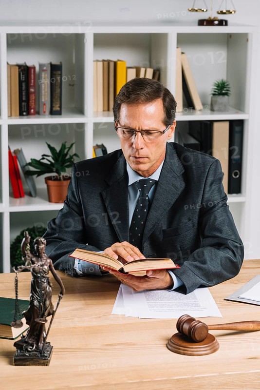 Mature male lawyer reading book with gavel justice statue wooden table