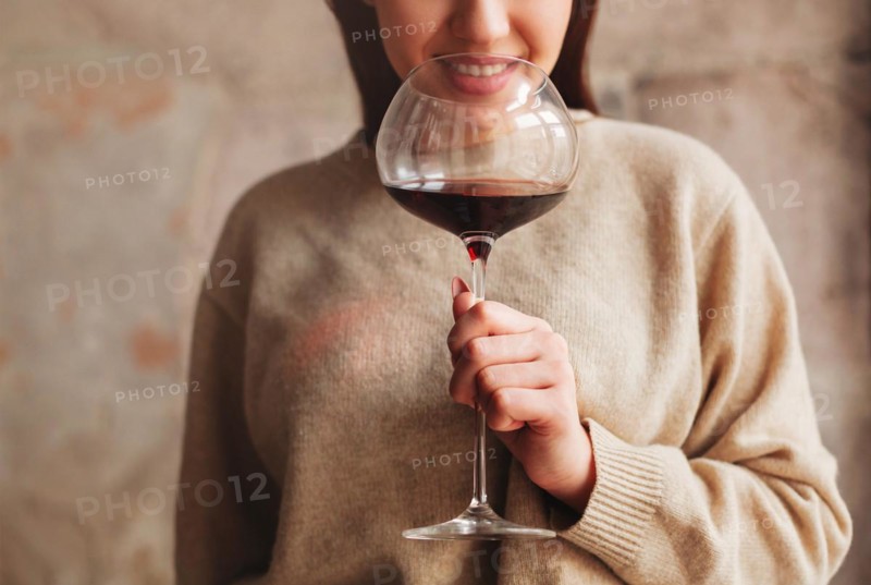 Crop happy female in sweater smelling red wine in large glass against gray background
