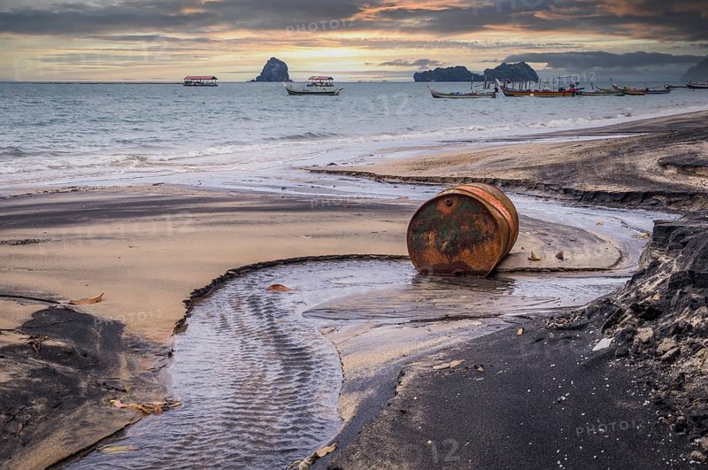 Rusty barrel oil on a partly black coloured beach illustrates the pollution of environment by oil spills. Longtail boats in the background at sunset