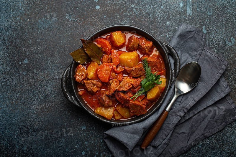 Beef meat stew with potatoes, carrot and delicious gravy in black casserole pot with bay leaves and fresh green herbs with spoon on black dark rustic concrete background from above, food photography, food photography