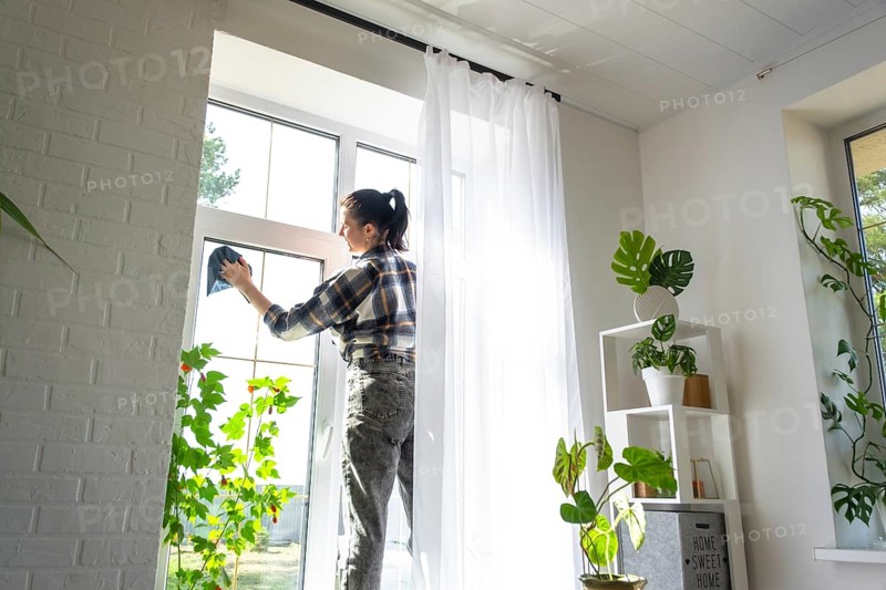 Woman manually washes the window of the house with a rag with spray cleaner and mop inside the interior with white curtains. Restoring order and cleanliness in the spring, cleaning servise