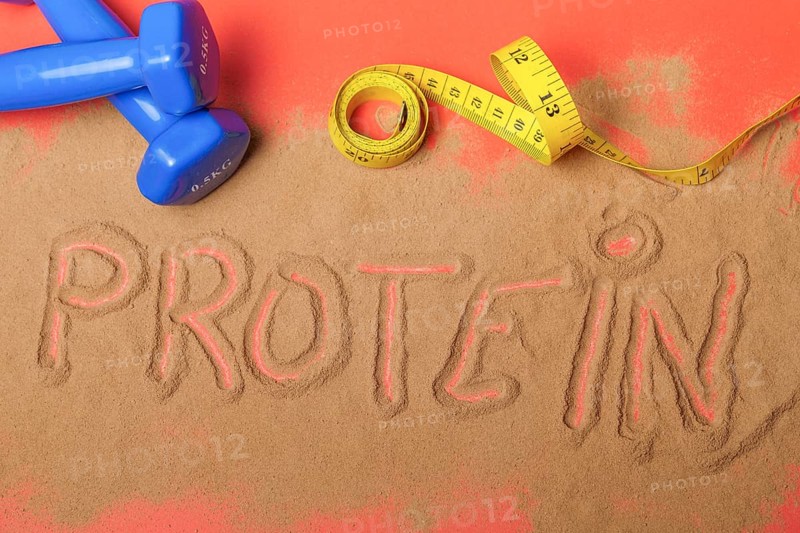 Word protein written on protein powder on red background, with dumbbells and measuring tape