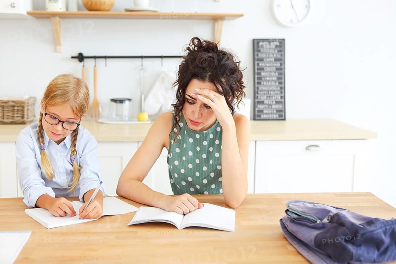 ?ute school girl wearing eyeglasses doing homework with mother at home on the kitchen
