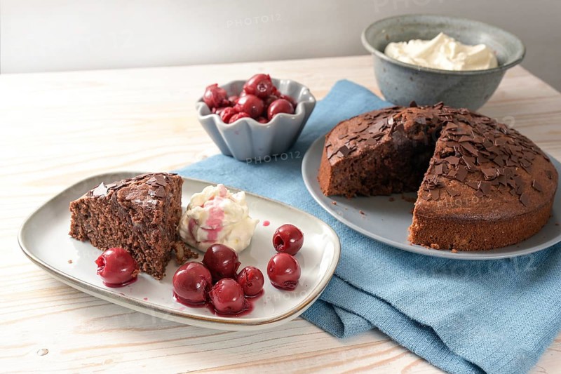 Homemade chocolate cake with morello cherries and whipped cream on a blue napkin and a bright wooden table, copy space, selected focus, narrow depth of field
