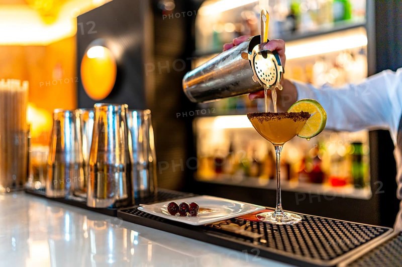 Hand of a waiter pouring cocktail into martini glass garnished with lemon in the counter of a luxury night club