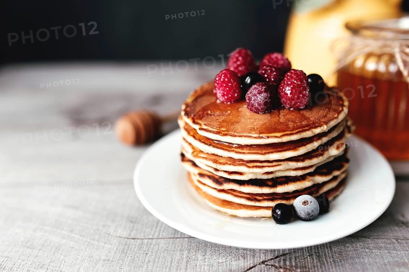 Juicy pancakes with berries and honey on a white plate, spoon, jar, wooden table, yellow vase with eucalyptus.