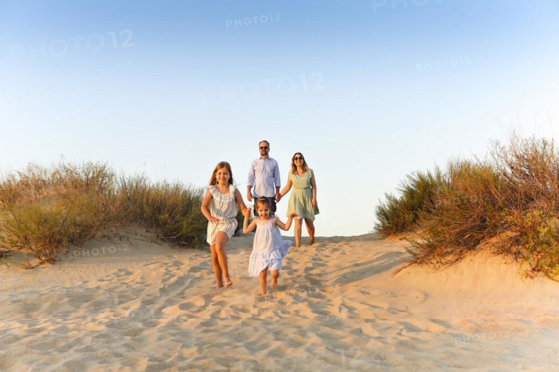 Happy family of four, young parents and little kids, enjoying summer vacation in nature, taking walk on sandy beach, holding each others hands, girls sisters running ahead while parents walking behind