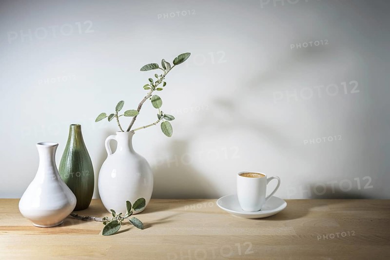 Pale still life with three vases in white and green, a sage leaf branch with shadow and a coffee cup, wooden table against a gray wall, take a break with simple home decor, copy space, selected focus