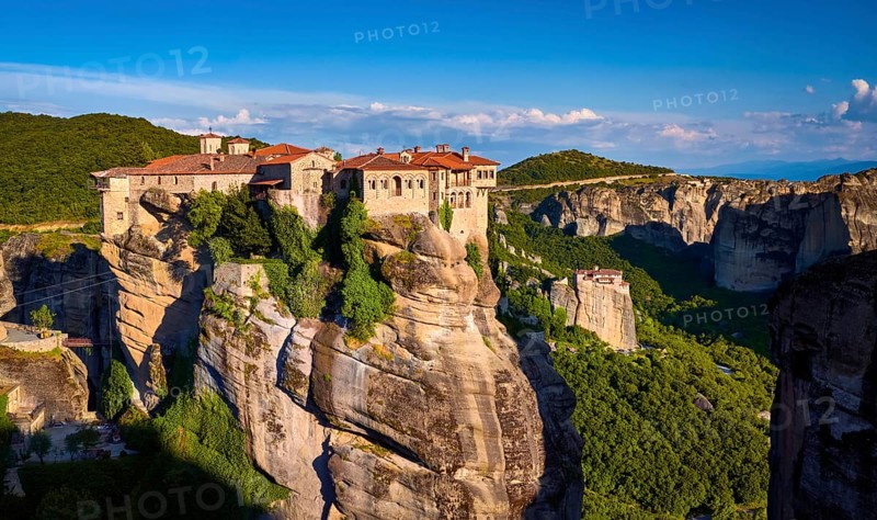 Clifftop Varlaam monastery lit by evening sunlight in Meteora, Greece in typical Meteora landscape of rocky pillars and slopes covered by rich spring foliage. Moni Agias Varvaras Roussanou nunnery in background. UNESCO World Heritage