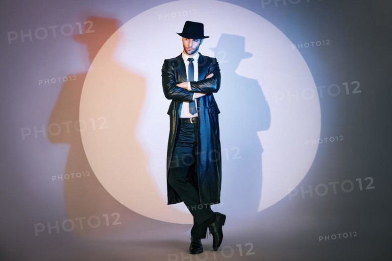 Full length portrait of confident handsome man in black coat and hat with crossed arms posing on studio in the spotlight. Noir film style. Private detective, spy, investigation concept