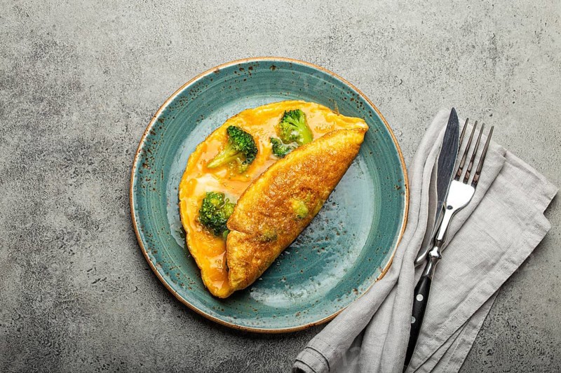 Healthy vegetarian egg omelette with green broccoli folded in half served on plate with fork and knife top view on gray stone concrete background table, diet and healthy clean eating concept