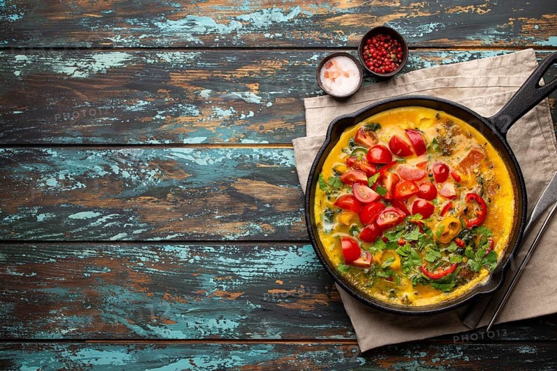 Healthy frittata in cast iron pan with fried beaten eggs and seasonal vegetables on rustic wooden background. Italian omelette with organic spinach, bell pepper, tomatoes, top view, copy space