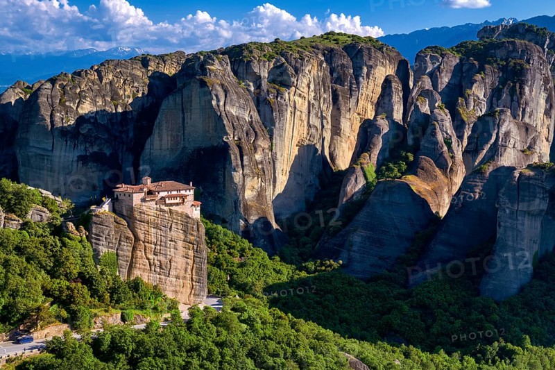 View of Meteora valley, Greece at midday. Clifftop Moni Agias Varvaras Roussanou nunnery among hills covered with bushes and trees and high sedimentary cliffs and rocks. Snow-covered mountains in distance. UNESCO World Heritage