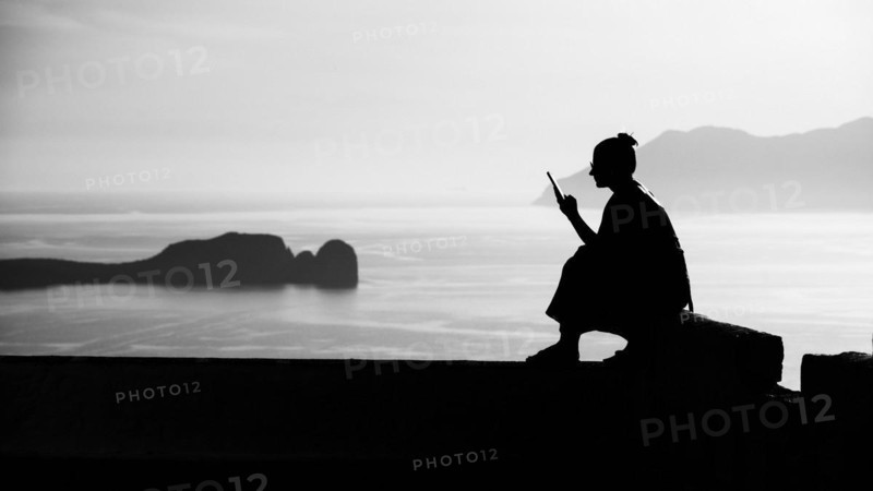 A woman sits on a stone wall and looks at her mobile phone, Venetian Castle, Plaka, Milos, Cyclades, Greece, Europe