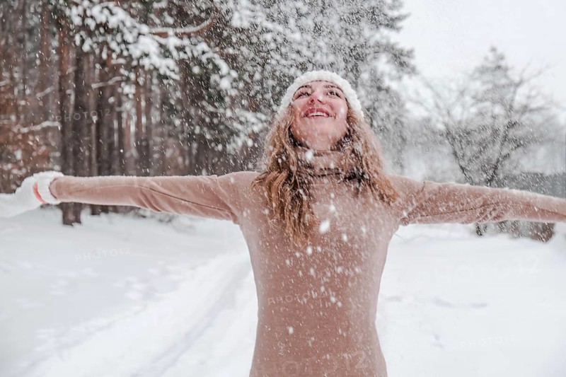 Christmas, holidays and season concept. Young happy woman blowing snow in the winter forest nature. Warm clothing knitted gloves and hat. Winter forest landscape background