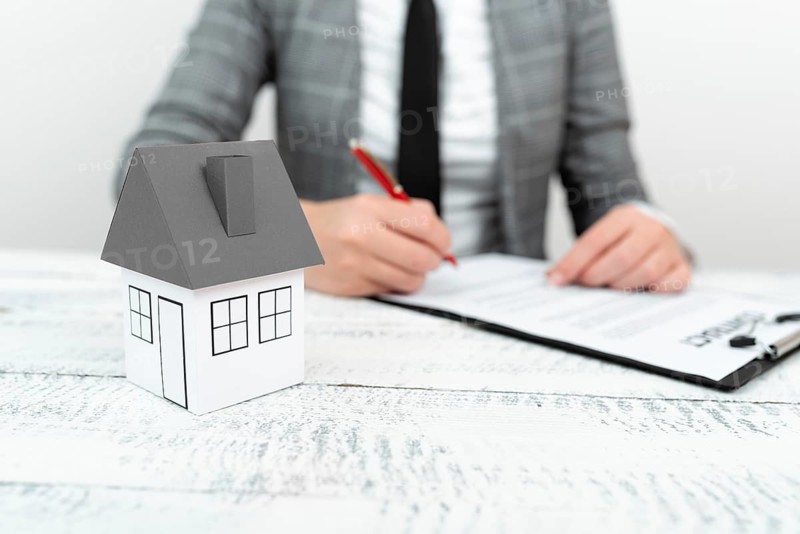 Businesswoman sitting and writing in notebook. Paper house on desk