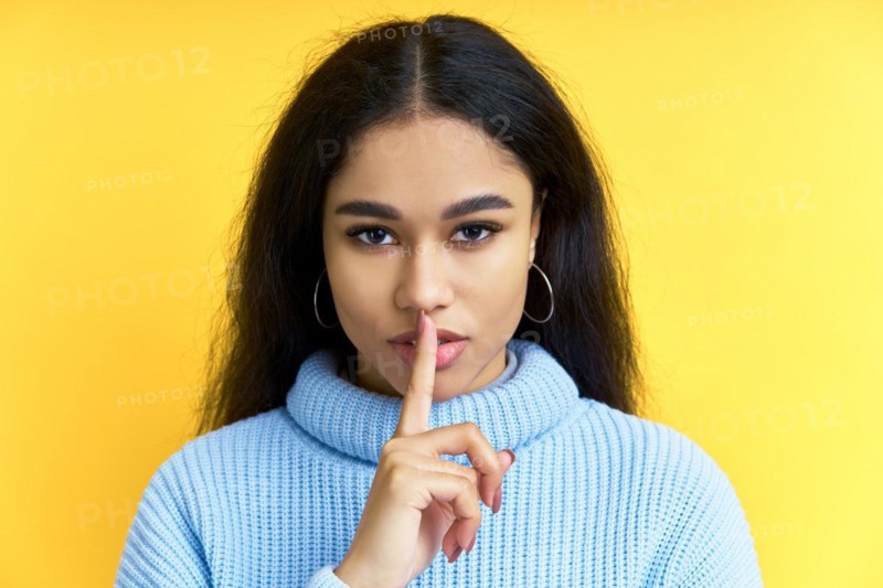 Pretty black woman keeps finger on lips, making hush gesture on yellow background. Shh, silence concept