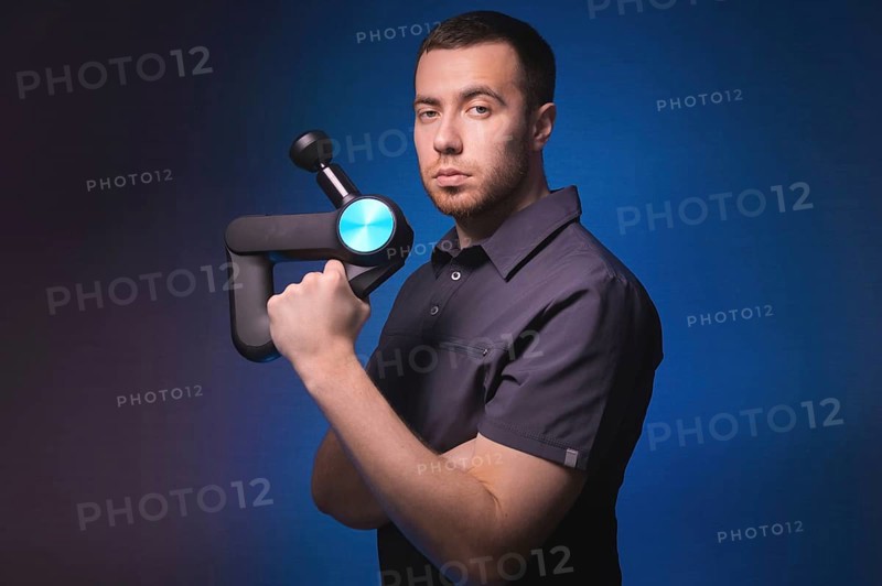Portrait of a confident professional masseur with a percussion massager in his hands. Low key, blue background. Shock wave massage