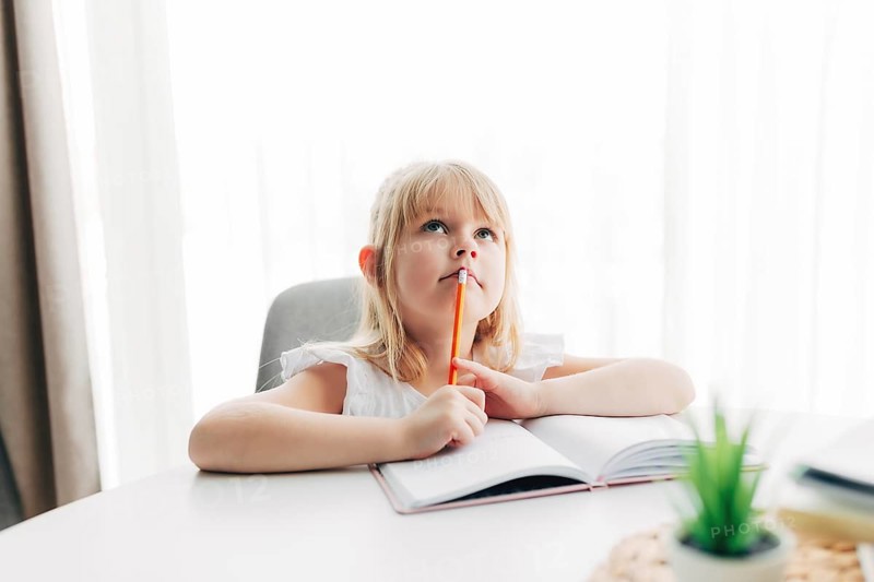 A little girl sits at the table and writes in a white notebook. Girl raised her finger, have an idea or got a result. Education concept. Home schooling. Homework. Girl is thinking on task solution