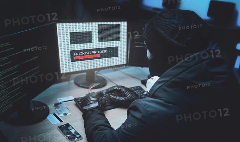 Shot from the Back to Hooded Hacker Breaking into Corporate Data Servers from His Underground Hideout. Place Has Dark Atmosphere, Multiple Displays