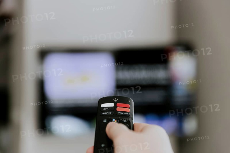Hand holding remote control pointing to TV. Person watching smart television indoors. Blurred background. Colorful screen, copy space. Person browsing, looking for films online at home living room