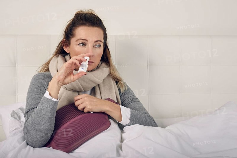 Sick woman wrapped in a warm scarf and clutching a hot water bottle sitting using nasal spray with a miserable expression in a seasonal healthcare or flu concept