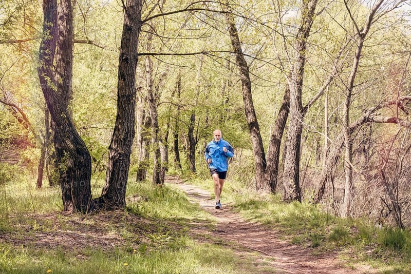 A senior man worn in black and blue is running in the forest, during a warm spring day