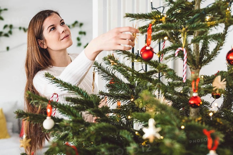 Beautiful smiling cheerful caucasian woman dressed in white outfit decorating a natural Christmas tree, putting red ornaments on the tree, holding a red Christmas ornament