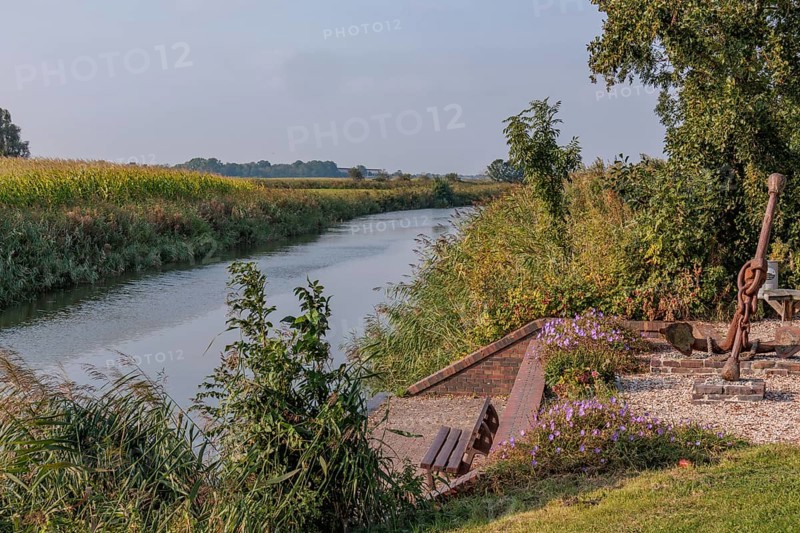 A quiet river flows along a bank with benches, flowers and trees, krummhörn, east frisia, germany